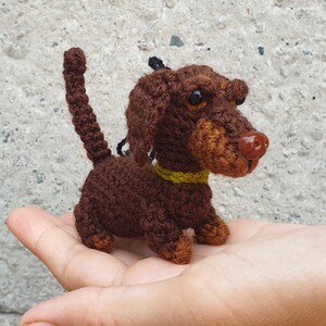 May include: A small brown crocheted dachshund with a yellow collar. The dachshund is sitting on a person's hand.