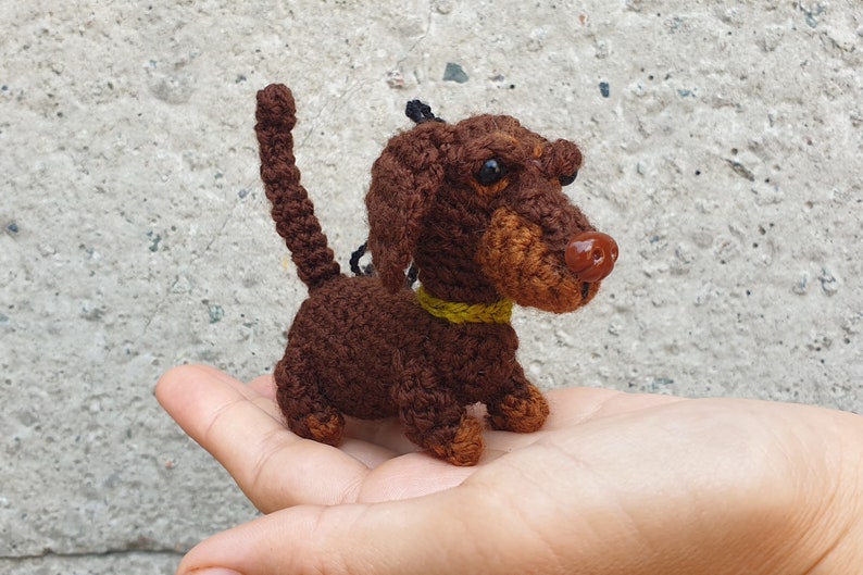 May include: A small brown crocheted dachshund with a yellow collar. The dachshund is sitting on a person's hand.