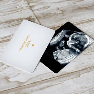 Puede incluir: Una tarjeta blanca con texto dorado que dice "Coming Soon. 2021." y un peque&ntilde;o coraz&oacute;n dorado. Una imagen de ultrasonido en blanco y negro est&aacute; encima de la tarjeta.