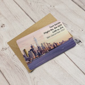 May include: A luggage tag featuring a cityscape of New York City at sunset. The tag has the text "Tom Smith, Flight No. BA 0221, LHR to JFK, 18th December 2021" printed on it.
