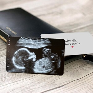 Puede incluir: Una cartera negra con una cremallera dorada y una tarjeta blanca con una imagen de ultrasonido en blanco y negro y el texto "Baby Alfie due 03.06.23" con un coraz&oacute;n rojo.