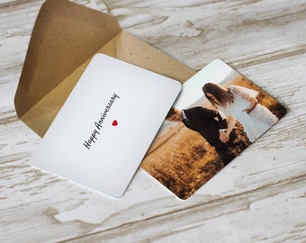 Puede incluir: Una tarjeta blanca con el texto "Happy Anniversary" y un pequeño corazón rojo. La tarjeta está junto a una foto de una pareja de pie en un campo.