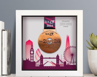 May include: A white shadow box frame containing a bronze 2024 TCS London Marathon finisher medal with a ribbon. The background features a pink and purple London skyline illustration with the text 27th April 2024, CHARLIE CADENCE, and 04:26:25.