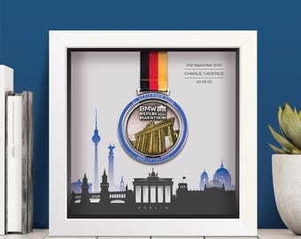 May include: A framed BMW Berlin Marathon medal with a black, red, and yellow ribbon. The medal is silver and blue, featuring the Brandenburg Gate. The frame includes a Berlin skyline silhouette and the text "21st September 2025".