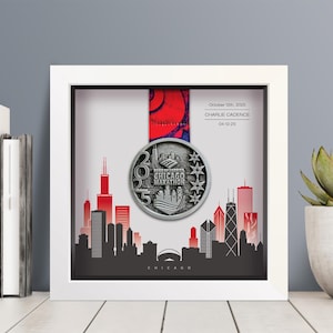 May include: A white shadow box frame containing a 2025 Chicago Marathon finisher medal suspended by a red and blue ribbon. The background features a black and red Chicago skyline graphic with the text October 12th, 2025, CHARLIE CADENCE, 04:12:25.