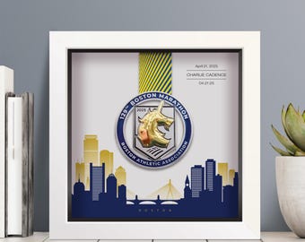 Może przedstawiać: Oprawiony medal z maratonu bostońskiego. Medal jest srebrny i złoty, z napisem "129th Boston Marathon 2025". Rama ma niebiesko-złotą wstążkę i sylwetkę panoramy Bostonu. Widoczny jest tekst "April 21, 2025" i "CHARLIE CADENCE".