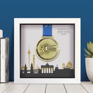 May include: A white shadow box frame containing a gold BMW Berlin Marathon medal with a blue ribbon. The background features a black silhouette of the Berlin skyline. Text reads 29th September 2024, CHARLIE CADENCE, and 03:26:25.