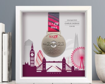 Może przedstawiać: Oprawiony medal ukończenia Maratonu Londyńskiego 2023. Srebrny medal z różowo-czarną wstążką, prezentowany w białej ramie. Tło przedstawia fioletową sylwetkę panoramy Londynu, w tym charakterystyczne budowle.