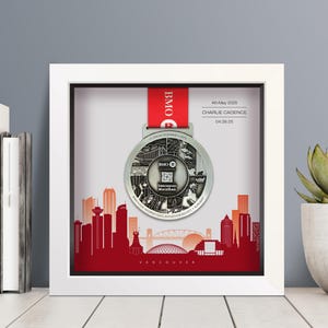 May include: A framed Vancouver Marathon medal with a red ribbon. The silver medal is displayed in a white shadow box, with a red skyline graphic at the bottom. Text on the medal and frame includes the date, name, and time of the race.