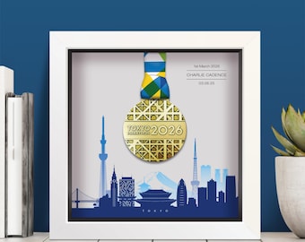 Może przedstawiać: Biała kwadratowa ramka typu shadow box zawierająca złoty medal Tokyo Marathon 2026 z geometrycznym wzorem, zawieszony na niebiesko-zielono-żółtej wstążce. Tło przedstawia niebieską sylwetkę panoramy Tokio oraz tekst: 1st March 2026, CHARLIE CADENCE, 03:26:25.