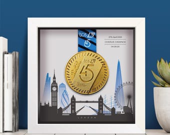 May include: A white square shadow box frame containing a gold TCS London Marathon medal suspended by a blue and black ribbon. The background features a black and blue London skyline illustration with the text 27th April 2025, CHARLIE CADENCE, 04:26:25.