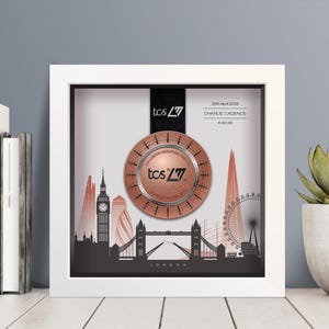 May include: A framed London Marathon medal with a cityscape silhouette. The medal is copper-colored and features the text "TCS London Marathon" and "26th April 2026". The frame is white, and the background is a light gray.