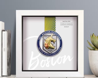 May include: A white shadow box frame containing a 129th Boston Marathon medal with a gold unicorn emblem, suspended by a yellow and blue striped ribbon. The background features the text April 21st, 2025, CHARLIE CADENCE, and 04:04:21.