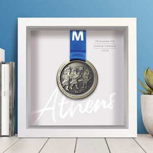 May include: A framed bronze medal with a blue ribbon. The medal depicts three figures in a scene from the Battle of Marathon. The text on the medal reads "Athens Marathon, The Authentic, Battle of Marathon 490 B.C.". The frame includes a white mount with the text "Athens" written in white script. The date "10th November 2019", the name "Charlie Cadence", and the time "4:37:28" are printed on the mount.