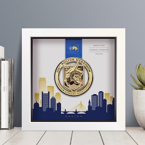May include: A framed Boston Marathon medal. The gold medal is displayed in a white shadow box with a blue ribbon. The frame includes a blue and gold skyline silhouette and the text "130th Boston Marathon". Additional text includes "April 20, 2026" and "Charlie Cadence".