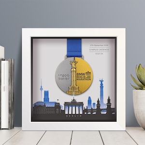 Pode incluir: Uma moldura quadrada branca tipo caixa contendo uma medalha da Maratona de Berlim BMW com uma fita azul. A medalha é metade prata e metade ouro, apresentando a Coluna da Vitória. Texto: 27th September 2026, Charlie Cadence, 03:27:26.