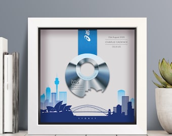 May include: A white shadow box frame containing a silver TCS Sydney Marathon medal suspended by a blue ribbon. The background features a blue silhouette of the Sydney skyline, including the Opera House and Harbour Bridge, with text reading 31st August 2025, CHARLIE CADENCE, and 03:31:25.