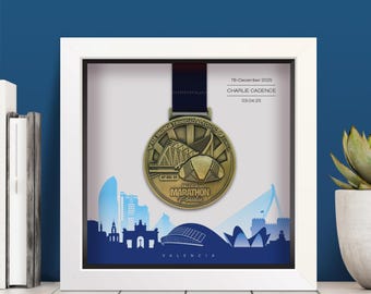May include: A white shadow box frame containing a gold-toned marathon medal suspended by a dark ribbon. The background features a blue silhouette of the Valencia skyline. Text reads: 7th December 2025, CHARLIE CADENCE, 03:04:25, and VALENCIA MARATHON.