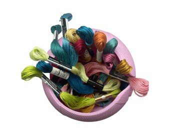 May include: A pink ceramic bowl filled with a variety of colorful embroidery floss spools. The spools are labeled "Cosmo" and have different numbers on them.