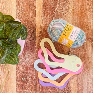 Puede incluir: Un ovillo de hilo de color azul claro, morado y blanco con una etiqueta amarilla que dice "REGIA desde 1949 BABY & Smiles My first REGIA". Hay cinco indicadores de tamaño de agujas de tejer de plástico de colores con forma de calcetín.