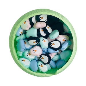 Puede incluir: Un bol verde lleno de cuentas de pingüino de silicona de colores. Las cuentas son en tonos de azul, rosa y negro.