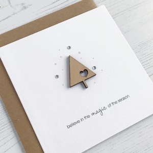 May include: A white greeting card with a wooden Christmas tree cutout featuring a heart-shaped cutout. The card has the phrase "believe in the magic of the season" printed in black. The card is on a brown envelope.