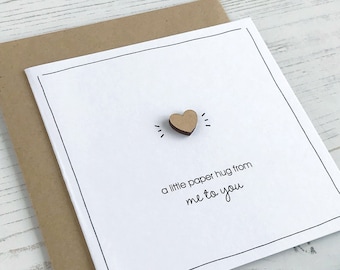 May include: A white card with a black border and the text "a little paper hug from me to you". A small wooden heart is attached to the card.