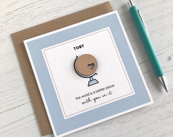May include: A greeting card with a light blue border and a brown envelope. The card features the name "TOBY" above a globe with a heart cutout. The text "the world is a better place with you in it" is below the globe. A teal pen rests on the side.