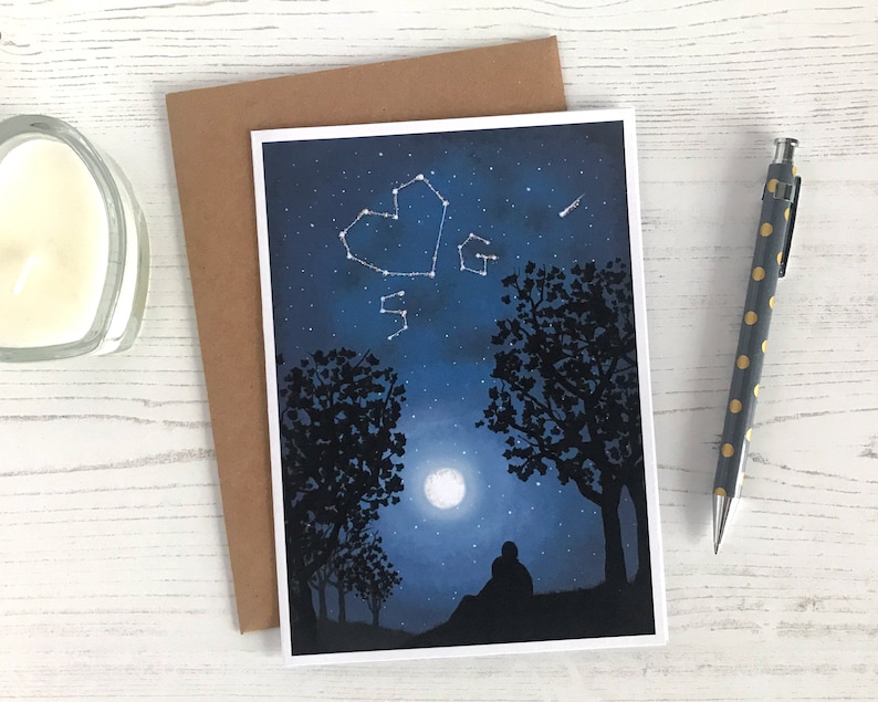 May include: A greeting card with a blue night sky background featuring a constellation in the shape of a heart, a full moon, and two silhouetted trees. The card has a white border.