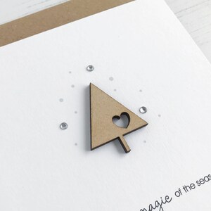 May include: A white greeting card with a wooden Christmas tree cutout featuring a heart. The card has small silver rhinestones and the phrase "magic of the season" printed in black.