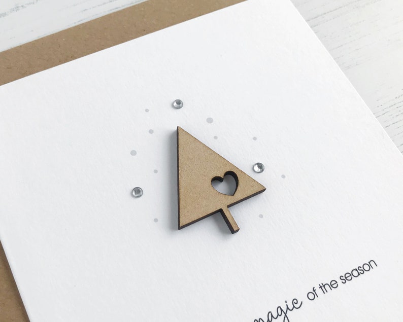 May include: A white greeting card with a wooden Christmas tree cutout featuring a heart. The card has small silver rhinestones and the phrase "magic of the season" printed in black.