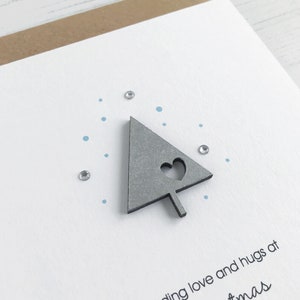 May include: A handmade Christmas card with a gray tree-shaped embellishment featuring a heart cutout. The card has a white background with blue dots and small, sparkling embellishments. The card is on a brown paper backing.
