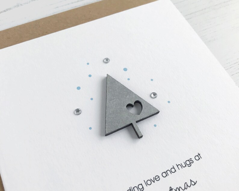 May include: A handmade Christmas card with a gray tree-shaped embellishment featuring a heart cutout. The card has a white background with blue dots and small, sparkling embellishments. The card is on a brown paper backing.
