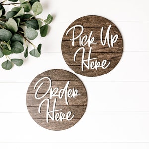 May include: Two round wooden signs with white script. The top sign says "Pick Up Here" and the bottom sign says "Order Here".