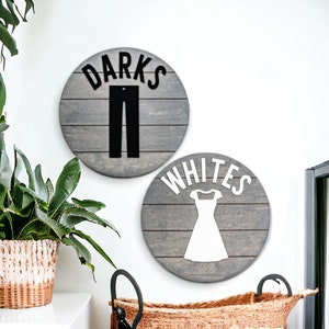 May include: Two round wooden signs with gray paint and white woodgrain. The sign on the left says "DARKS" with a black silhouette of pants. The sign on the right says "WHITES" with a white silhouette of a dress.