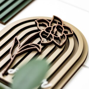 Op de afbeelding: Houten wandkunst met een gelaagd ontwerp en een bloemmotief. Het kunstwerk toont een narcis-silhouet in donkerbruin, tegen een lichtbeige gebogen achtergrond met zwarte strepen. Geschikt voor interieurdecoratie.
