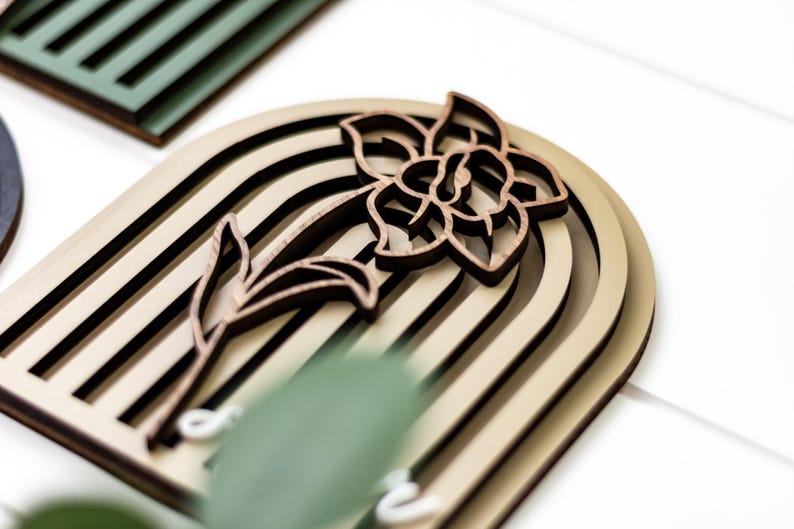 Op de afbeelding: Houten wandkunst met een gelaagd ontwerp en een bloemmotief. Het kunstwerk toont een narcis-silhouet in donkerbruin, tegen een lichtbeige gebogen achtergrond met zwarte strepen. Geschikt voor interieurdecoratie.