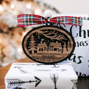May include: An oval wooden Christmas ornament featuring a laser-cut winter village scene with pine trees and houses. The text reads, "THERE'S NO PLACE LIKE 30180 HOME FOR THE HOLIDAYS." It is topped with a white bead and a red, green, and blue plaid ribbon.
