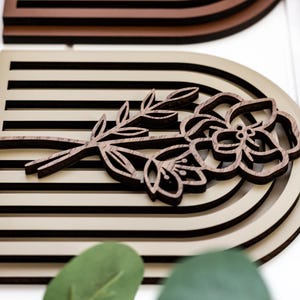 May include: A wooden wall decor piece featuring a layered, arched design in beige and brown tones. The arch is decorated with a carved wooden flower and stem in a dark brown colour.