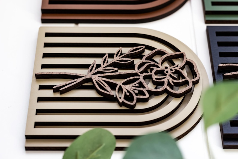 May include: A wooden wall decor piece featuring a layered, arched design in beige and brown tones. The arch is decorated with a carved wooden flower and stem in a dark brown colour.