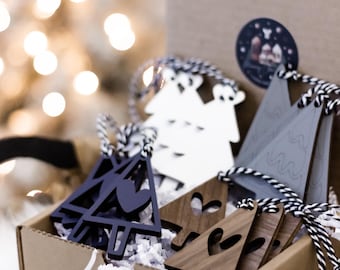 May include: A cardboard box filled with wooden Christmas tree ornaments in various colors, including navy, white, gray, and brown. The ornaments have heart and tree cutouts and are tied with black and white twine. The box is on a white fluffy surface.