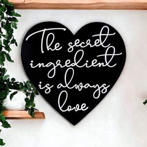 May include: Black heart-shaped sign with white text that reads "The secret ingredient is always love".