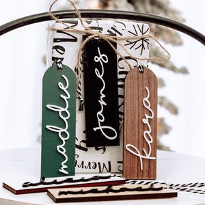 May include: Three wooden gift tags with white lettering. The tags are green, black, and brown. The names "Maddie", "James", and "Kara" are written on the tags.