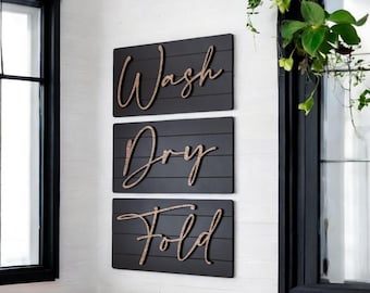 May include: Three rectangular black wooden signs with the words "Wash," "Dry," and "Fold" in a cursive font. The signs are mounted on a white wall, with a window and greenery visible in the background. The signs are a decorative laundry room theme.