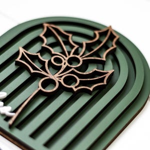 Op de afbeelding: Een houten kerstversiering met een hulstbladontwerp. De versiering heeft een donkergroene gewelfde achtergrond met een gelaagd ontwerp. Het hulstblad is bruin en gedetailleerd. Het woord "little" is zichtbaar in het wit.