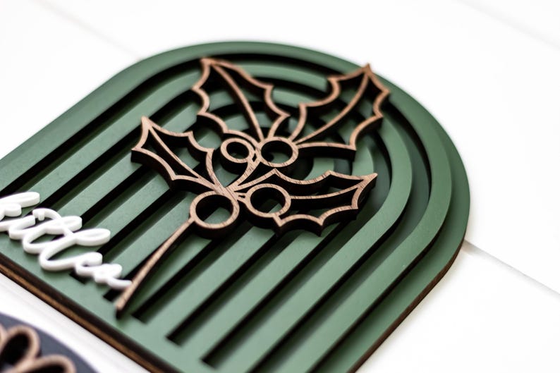 Op de afbeelding: Een houten kerstversiering met een hulstbladontwerp. De versiering heeft een donkergroene gewelfde achtergrond met een gelaagd ontwerp. Het hulstblad is bruin en gedetailleerd. Het woord "little" is zichtbaar in het wit.