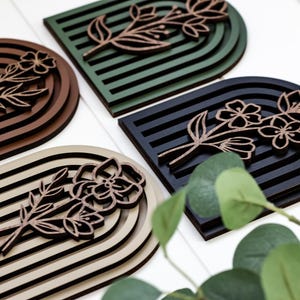 May include: Four wooden wall decor pieces in different colours, each featuring a floral design with a layered arch shape. The colours are brown, green, black, and beige.