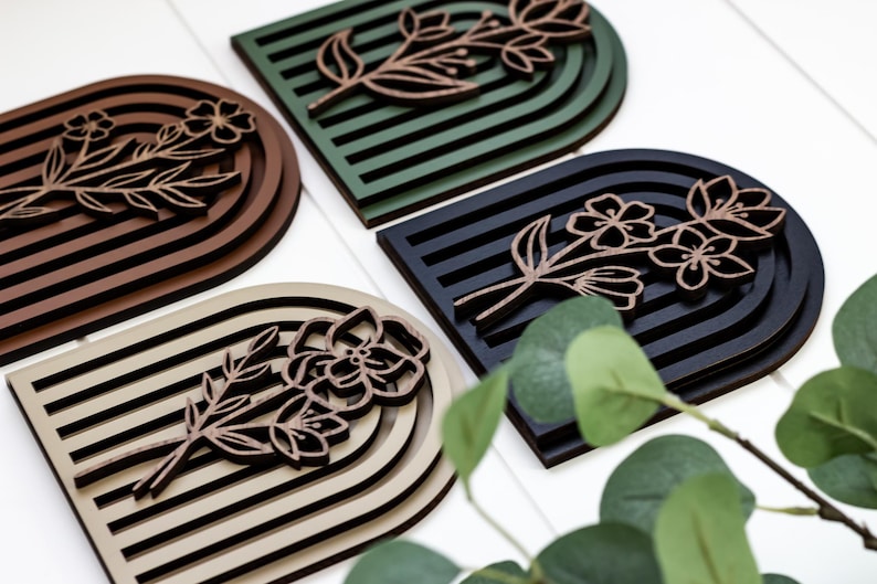 May include: Four wooden wall decor pieces in different colours, each featuring a floral design with a layered arch shape. The colours are brown, green, black, and beige.