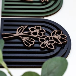 May include: A black and brown wooden wall decoration with a floral design. The design features a branch with three flowers and leaves. The background is a black and green striped pattern.