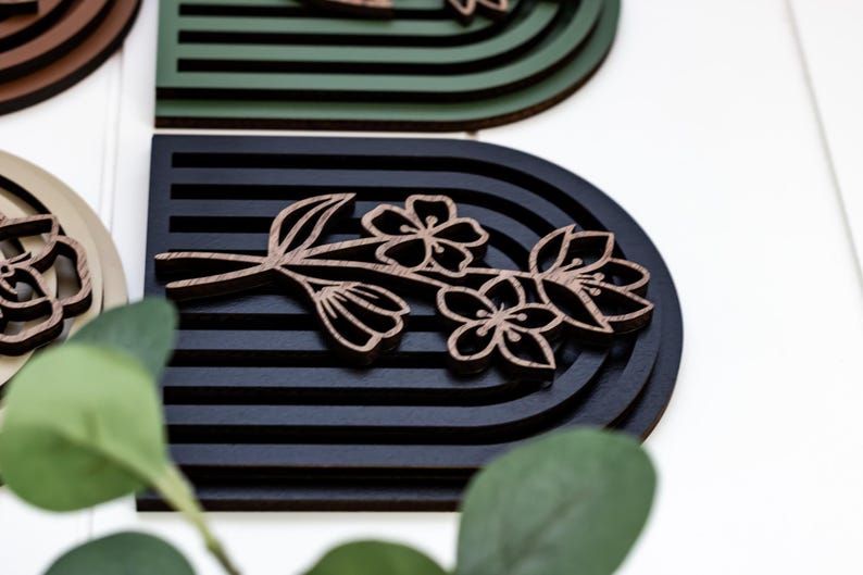 May include: A black and brown wooden wall decoration with a floral design. The design features a branch with three flowers and leaves. The background is a black and green striped pattern.